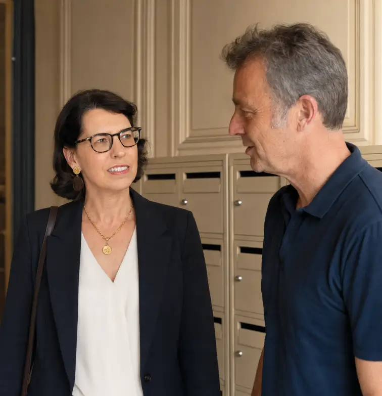 Une gestionnaire du cabinet d'administration de biens Warren & Associés échange de manière bienveillante avec un gardien d'immeuble devant les boîtes aux lettres de la copropriété.